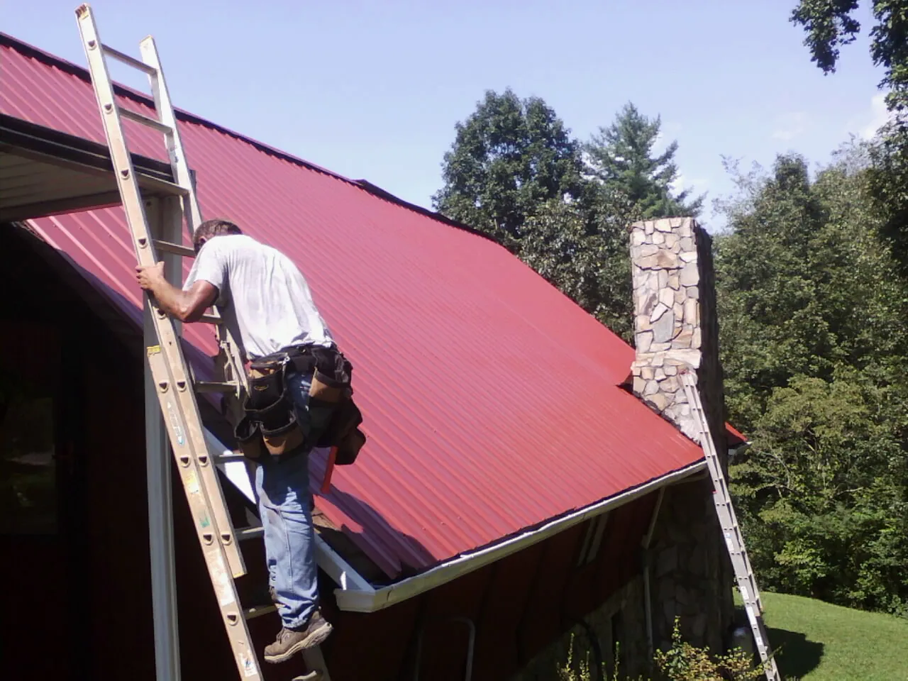 Roofer on a ladder beside a red metal roof and stone chimney — Soffit & Fascia Repair by Roofing Brothers in Black Diamond